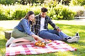 Happy diverse couple sitting on blanket and having picnic in sunny garden, copy space