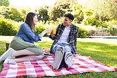 Happy diverse couple sitting on blanket, having picnic, making a toast in sunny garden, copy space