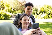 Happy diverse couple sitting and using smartphone in sunny garden, copy space