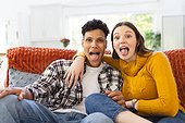 Portrait of happy diverse couple sitting on couch, embracing, making funny faces at home, copy space