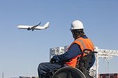 Transportation engineer in wheelchair watching airliner flying over shipping containers and cranes at port